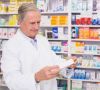 Älterer Mann im Kittel steht in Apotheke mit Regalen voller Medikamente hinter sich und hält Pappschachtel in der Hand