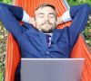 Happy businessman with laptop relaxs in a hammock on nature. Fre